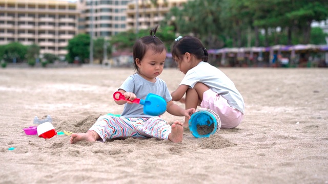 一个小女孩和她的小妹妹在沙滩上玩沙子，后面是城市的景色视频素材