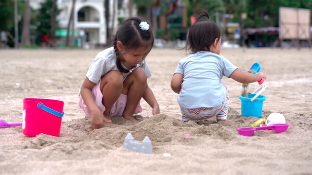 一个小女孩和她的小妹妹在沙滩上玩沙子，后面是城市的景色视频素材