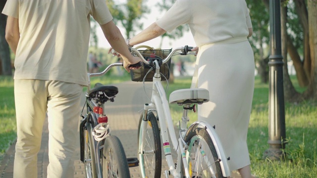 后视图，不认识的中年男人和女人走在自行车在夏季公园日落。一对活跃的白种夫妇在外面的巷子里散步。永恒的爱,成键。视频素材