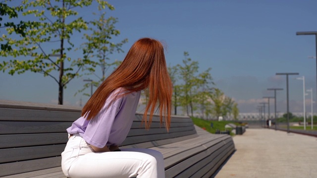 在阳光明媚的日子里，一个漂亮的红头发女孩穿着休闲衣服坐在城市公园的长椅上。视频素材