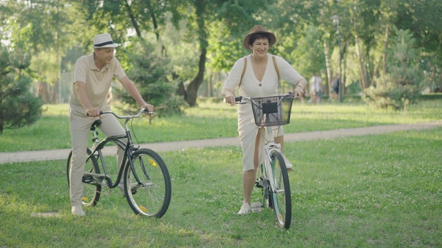 欢乐的中年白人夫妇在阳光明媚的夏季公园骑自行车的肖像。丈夫跟着可爱的妻子骑着自行车离开。快乐的家庭享受户外爱好。键,爱。视频素材