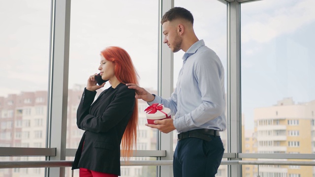 男人给红头发的女人礼物。一个打电话的女人从她的丈夫那里收到了一份礼物，并且在办公室里很高兴收到这份礼物。视频素材