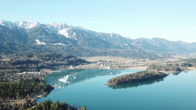 奥地利Faaker See上空的一架无人机。周围有高山山脉，反射在清澈平静的湖面上。小村庄围绕着湖。白雪皑皑的山脉视频素材