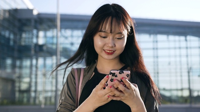 愉快的无忧无虑的年轻亚洲女人打字短信在她的手机附近的机场大楼视频素材