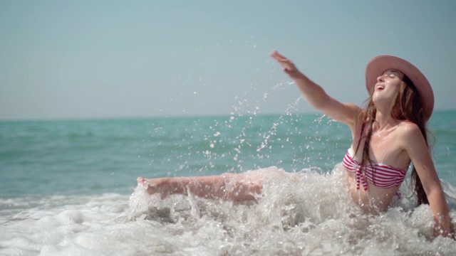 温暖汹涌的海浪拍打着坐在海边戴着夏日帽子的漂亮女孩。快乐的白人女人在性感的比基尼享受暑假，躺在水和玩海浪。无忧无虑的视频素材