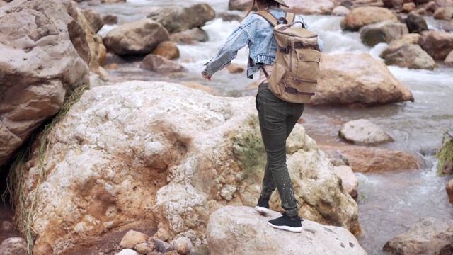 年轻美丽的亚洲女人背包徒步旅行和跳在岩石上的小溪在暑假。快乐漂亮的女孩，放松和享受美丽的自然。旅行和户外活动的概念视频素材