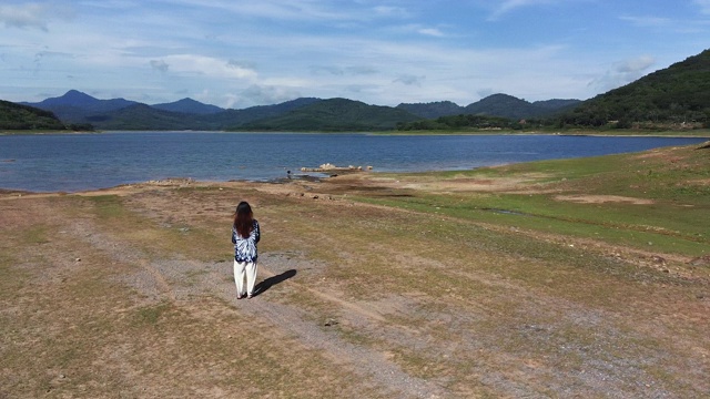 一个女人站在岩石地面上看一个湖和山的景观在一个阳光明媚的日子与一些云。她的头发在动。复制空间的旗帜，文本。视频下载