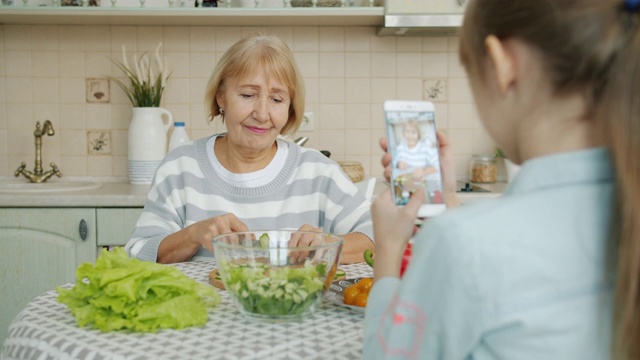 小女孩在拍奶奶做沙拉的照片，然后拿着黄瓜片摆姿势拍照视频素材