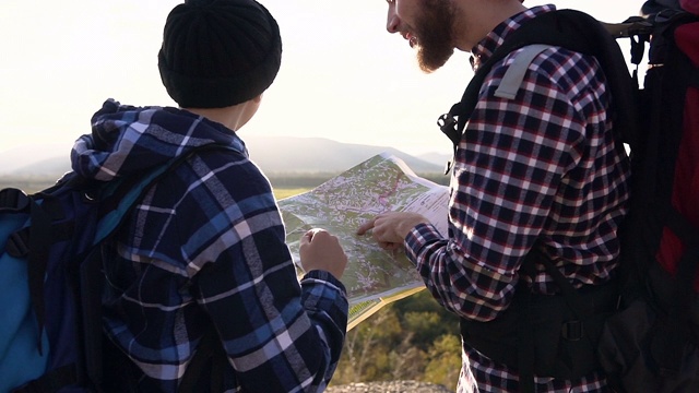 后视图。特写的年轻夫妇与背包和旅游地图，站在山顶享受徒步旅行的山，试图找到方向视频素材