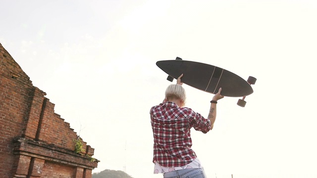 后视图，女孩穿着格子衬衫和牛仔裤与滑板在她的手是站在屋顶现代滑板公园在夏季时间日落。一个年轻女子手拿着长长的滑板站在那里，看着这座城市视频素材