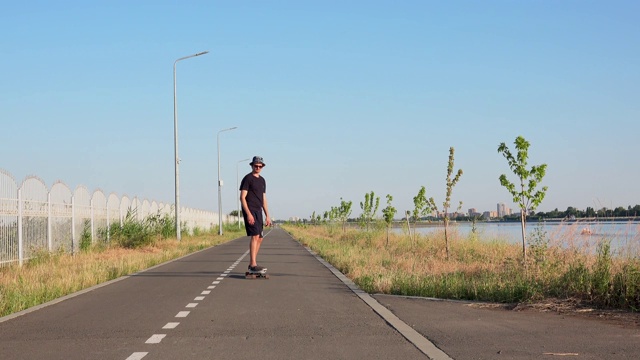 男子骑着长板在江边的空直路。夏日潮人在帽子骑滑板在城市外的日落时间4k视频素材