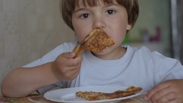 孩子吃披萨。饥饿的小男孩坐在家里厨房的桌子上吃着美味的意大利披萨。不健康的营养快餐。一个吃午饭的孩子的肖像。面对小男孩特写。视频素材