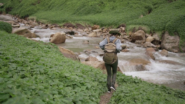 慢镜头后视图年轻美丽的亚洲女人背包徒步旅行在山小径附近的河流在暑假。美丽的女孩放松，享受与美丽的自然散步。旅行和户外活动的概念。视频素材