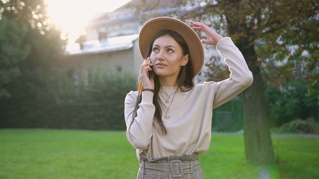 微笑着漂亮的年轻女子在米色帽子站在附近的绿树在房子的院子里和打电话视频素材