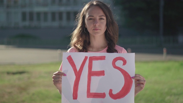 一个年轻的黑发女人在户外拿着Yes的海报。严肃的白人女性活动家举着标语站在阳光下。漂亮的抗议者在一个人的抗议中。视频素材