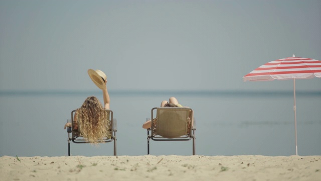 两个美丽放松的年轻女子躺在日光浴躺椅和日光浴。在阳光明媚的避暑胜地休息的快乐的白人女性游客的后视图宽镜头。放松的概念。视频素材