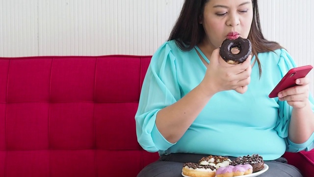 大码亚洲女性喜欢吃甜甜圈和在家里使用智能手机视频素材