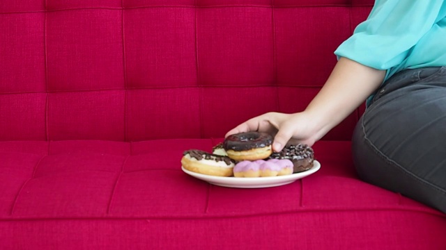 近手加大尺寸的女人喜欢坐在房间的红色沙发上吃甜甜圈。手选甜甜圈吃视频素材