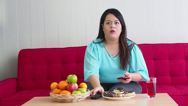 胖女人喜欢坐在房间的红色沙发上吃甜甜圈。一名女子一边用电视遥控器和手机，一边在客厅的桌子上放水果和甜甜圈。视频素材