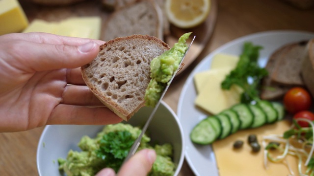 俯视图将牛油果泥抹在一片全麦面包上。健康的有机素食早餐在乡村风格。白色盘子，背景是纯素奶酪和蔬菜视频素材