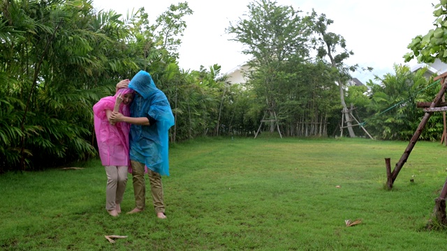 幸福的情侣在雨中穿着粉色和蓝色的雨衣在后院打转。视频素材