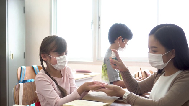 年轻女教师在教室里用酒精喷雾器消毒学生的手。亚洲妇女戴口罩，用洗手液清洁学生的手。学校在隔离和封锁后重新开学视频素材