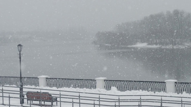 雪落在空旷的城市公园。湖边孤零零地立着一张白雪覆盖的长凳视频下载