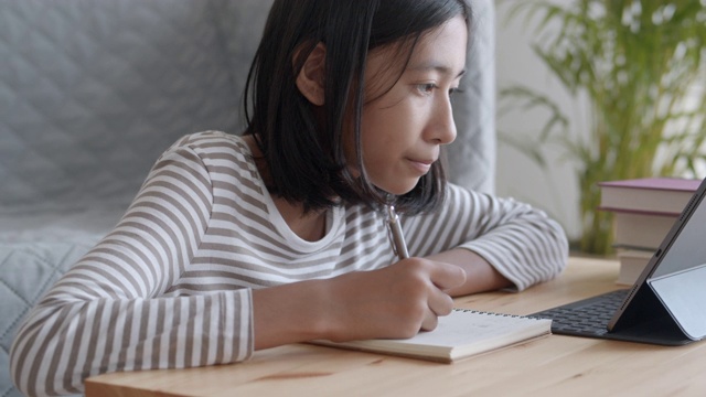 手持。一个亚洲女孩正在做作业，早上在桌子上用平板电脑。在家在线学习概念视频素材