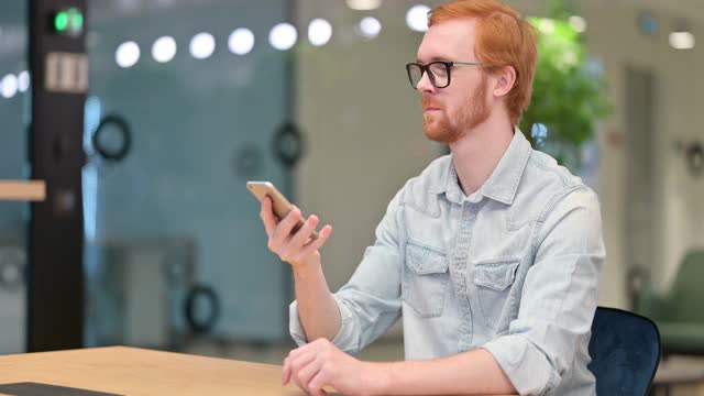 年轻的红发男子在办公室用智能手机聊天视频素材