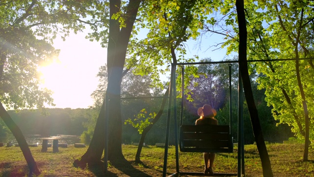 在金色的夏日夕阳下荡秋千的女人。戴草帽的快乐少女坐在秋千上打滚，欣赏美丽的大自然视频素材