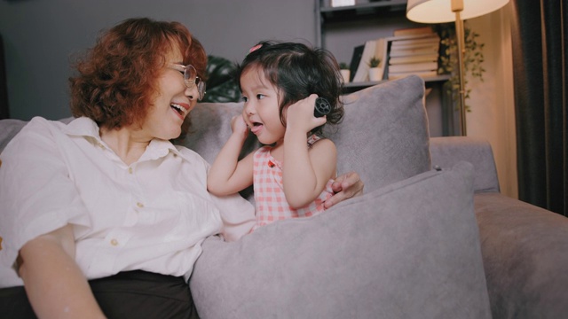 迷人的亚洲老祖母微笑着和孙子孙女在家里的客厅里玩，老女性开心的同时照顾着她可爱的小孩子。视频素材