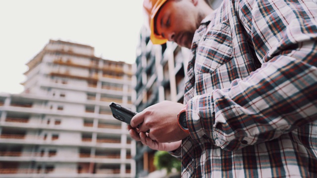 戴着防护头盔的白人男性建筑工人在正在建造的建筑附近使用智能手机。工程师检查施工过程，在电话上做记录。领班聊天应用视频素材