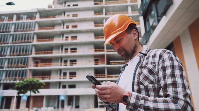 戴着防护头盔的白人男性建筑工人在正在建造的建筑附近使用智能手机。工程师检查施工过程，在电话上做记录。领班聊天应用视频素材