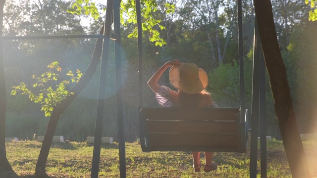 在金色的夏日夕阳下荡秋千的女人。戴草帽的快乐少女坐在秋千上打滚，欣赏美丽的大自然视频素材