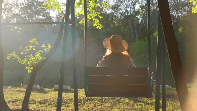 在金色的夏日夕阳下荡秋千的女人。戴草帽的快乐少女坐在秋千上打滚，欣赏美丽的大自然视频素材