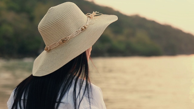 迷人的亚洲女人戴着软呢帽站在海滩上看海景美丽的日落。视频素材