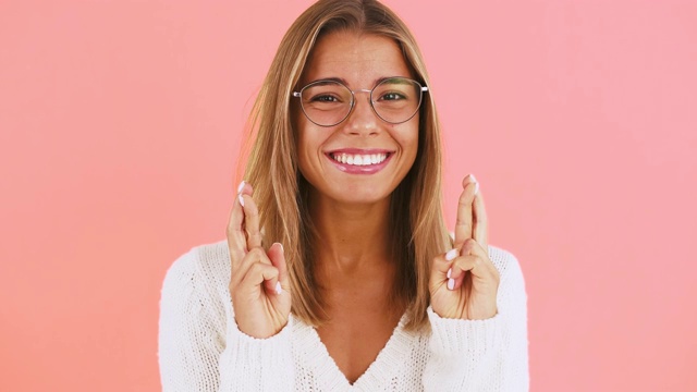 戴眼镜的年轻女士正在交叉她的手指，许愿，说请和在粉红色的背景下摆姿势微笑视频素材