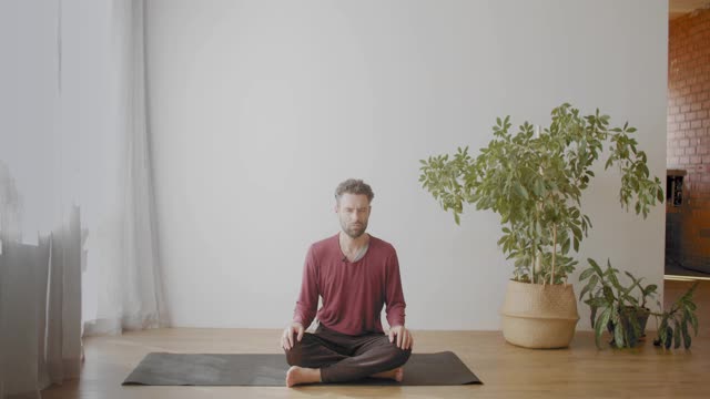 在阳光明媚的公寓里，一个白人男子盘腿坐着，闭着眼睛沉思。肖像的胡子瑜伽教练放松在苏哈沙那体式慢动作。放松压力克服视频素材