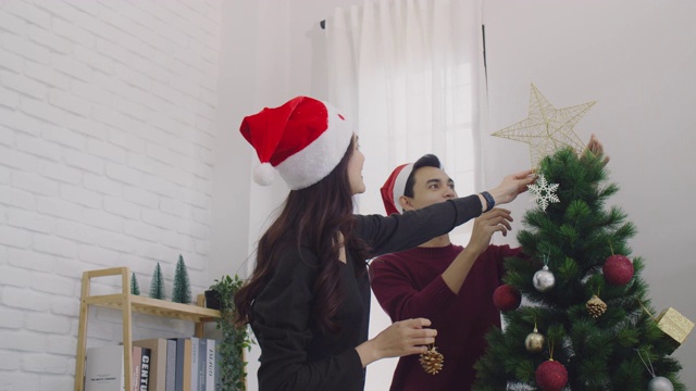 年轻的亚洲夫妇装饰圣诞树在圣诞节和快乐的新年节日在家里的背景。圣诞节庆祝情人的概念。4 k慢动作。视频素材