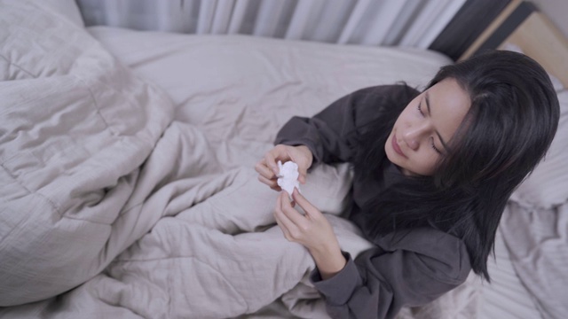 上面拍摄的年轻亚洲女人穿着毛衣坐在床里面的毯子打喷嚏，女人感觉不舒服在卧室里视频素材