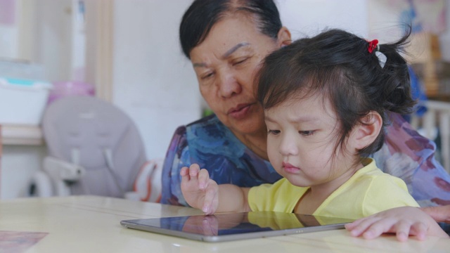 手持POV拍摄奶奶和孙子在家客厅看数字平板电脑。快乐的爷爷和孙女一起看电子屏幕，在家里放松。可爱的小女孩用手指触摸屏幕。视频素材