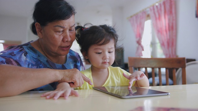 手持POV拍摄奶奶和孙子在家客厅看数字平板电脑。快乐的爷爷和孙女一起看电子屏幕，在家里放松。可爱的小女孩用手指触摸屏幕。视频素材