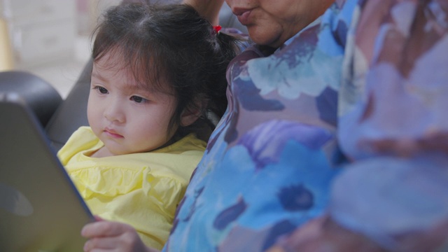 手持POV拍摄奶奶和孙子在家客厅看数字平板电脑。快乐的爷爷和孙女一起看电子屏幕，在家里放松。可爱的小女孩用手指触摸屏幕。视频素材