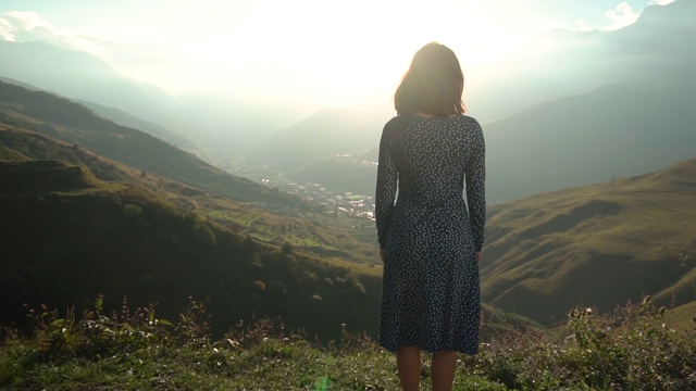 一个穿着裙子的年轻女子站在那里，举起双手望着群山。太阳从山的一侧明亮地照耀着。缓慢的运动。视频素材