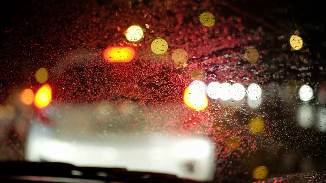 雨滴落在汽车挡风玻璃上。车内一辆车在行驶，模糊的红绿灯背景虚化。关闭了。视频素材