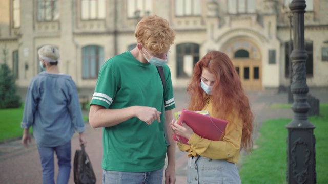 戴着口罩的大学生们集中在户外检查作业，模糊的男男女女在背景中互相问候。2019冠状病毒病大流行教育和生活方式。视频素材