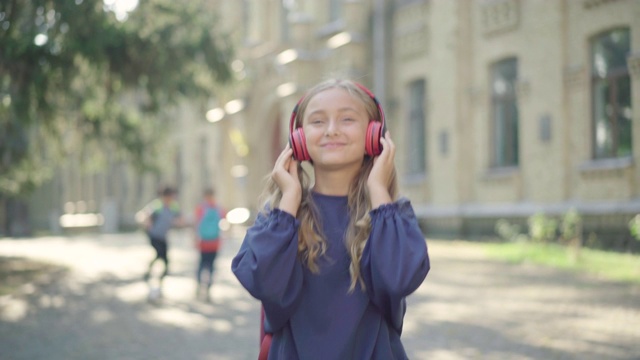 一个快乐的小女孩戴着耳机在阳光明媚的校园里听音乐。迷人的白人女学生享受夏日户外学习后。教育休闲理念。视频素材