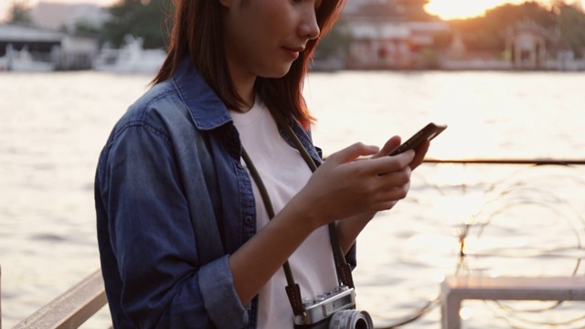 美丽的亚洲女人使用智能手机发送信息给她的朋友，而站在湄南河在美丽的日落。视频素材