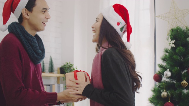 年轻的亚洲夫妇给对方一个礼物盒在圣诞节和新年期间在家里的背景与圣诞树装饰节日节日。圣诞节庆祝活动的概念。4 k慢动作。视频素材