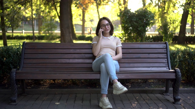 一个戴眼镜的年轻女子坐在长凳上讲电话。在背景中自然。视频素材